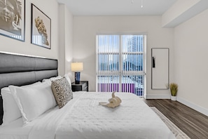 A modern hotel room with a large bed, a nightstand, a lamp, a mirror, and a window with a view of buildings. - Edgewater 2BR | Pool + Free Parking (Miami)
