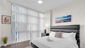 Une chambre d’hôtel moderne avec un grand lit, une table de chevet, une lampe, un tableau et une vue à travers des rideaux transparents.