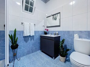 Un baño moderno con un mueble oscuro, lavamanos blanco y retrete. Hay dos plantas en macetas y un espejo sobre el lavamanos.