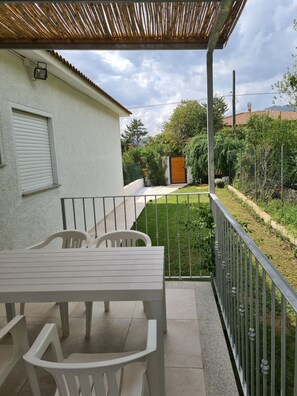 Un porche cubierto con una mesa y sillas, un jardín cercado y un edificio con techo de tejas al fondo.