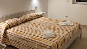 Una cama bien hecha con ropa de cama a rayas y una toalla doblada, una mesita de noche y una lámpara fijada a la pared.
