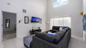 A modern living room with a grey sofa, a flat-screen TV, and a large window with curtains. - Disney Vila, Near Parks, Private Pool & Arcade (Kissimmee)