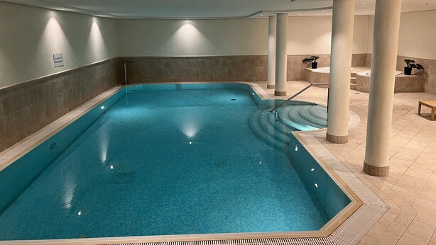 Un'ampia piscina coperta con acqua cristallina e azzurra, circondata da piastrelle color beige e colonne, con una panchina vicino al bordo.
