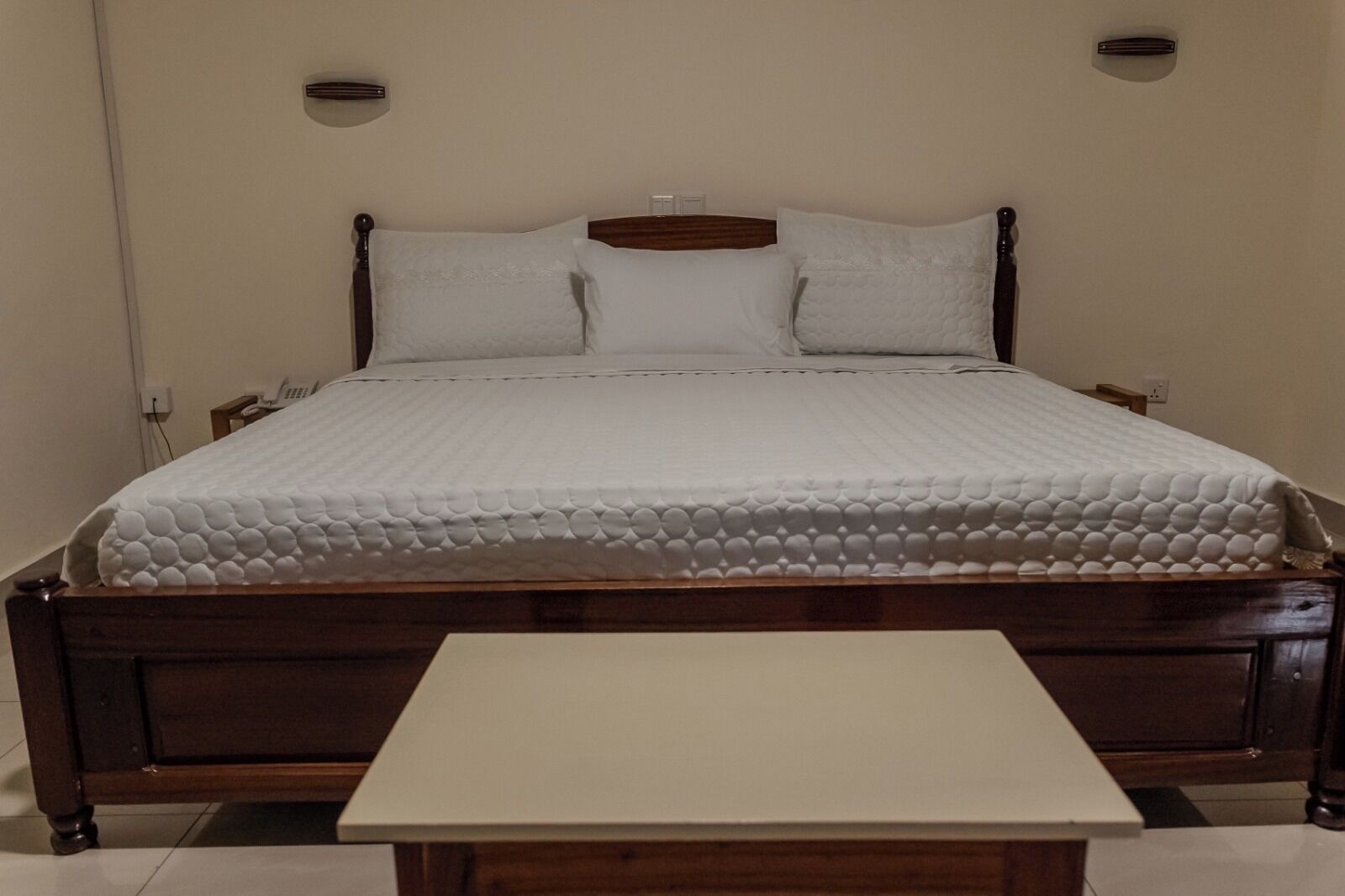 A bed with a wooden frame and white bedding, a bedside table, and a wall-mounted light fixture.