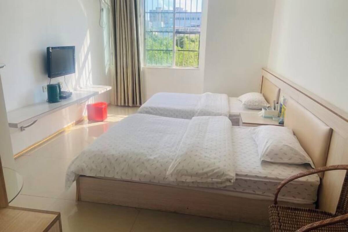 A hotel room with two beds, a TV on a shelf, a window with curtains, and a view of greenery outside.