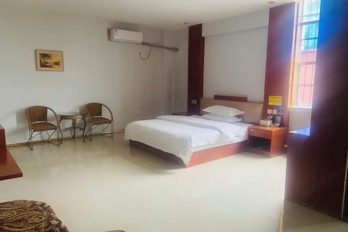 A hotel room with a bed, two chairs, a small table, a nightstand, an air conditioning unit, and a window.