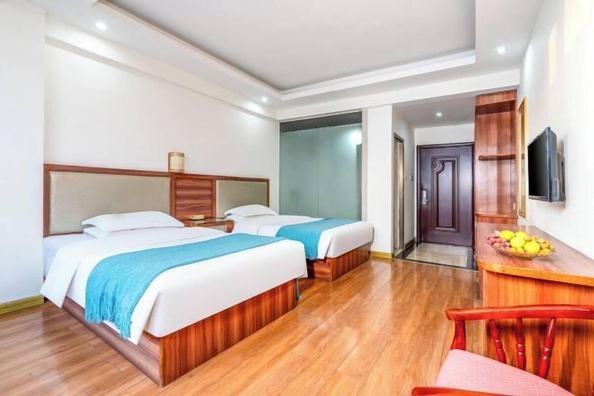 Ein Hotelzimmer mit zwei Betten, einem Holztisch mit Obstschale, einem Fernseher und einer Tür zu einem weiteren Raum.