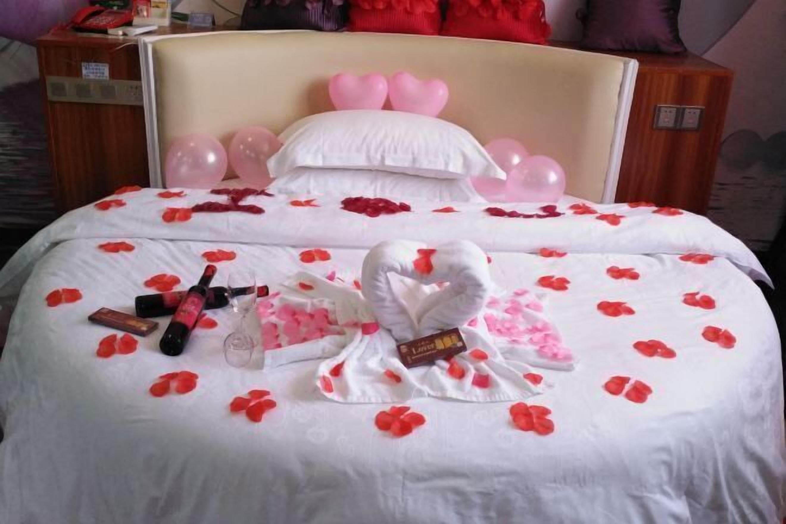 A hotel room with a bed adorned with rose petals, a heart-shaped towel, and pink balloons.