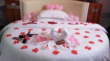 A hotel room with a bed adorned with rose petals, a heart-shaped towel, and pink balloons.