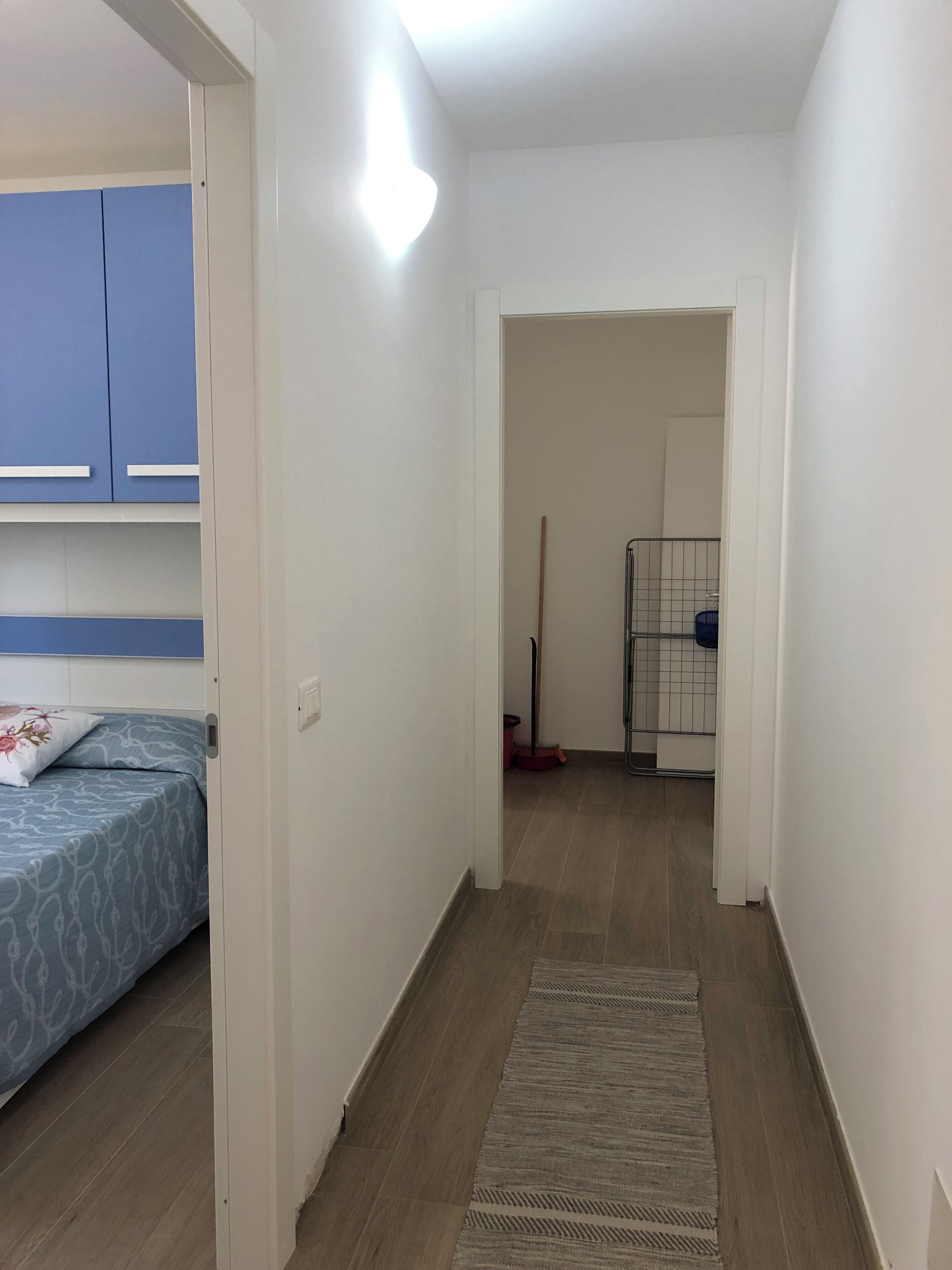 Un pasillo estrecho con una cama a la izquierda, una puerta al final y una escoba en la distancia.