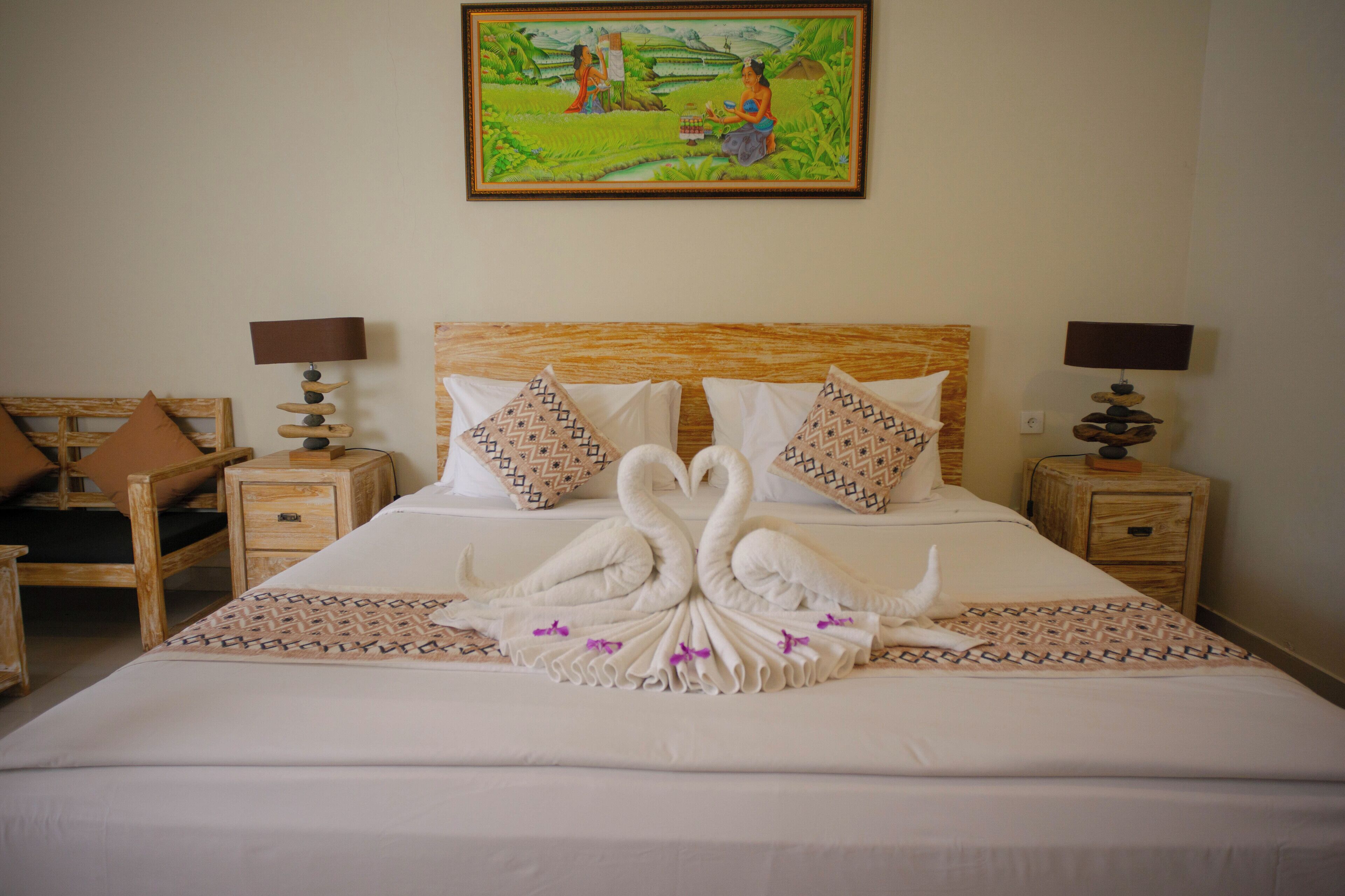 A neatly made bed with a swan towel design, two bedside tables with lamps, a wooden headboard, and a painting on the wall.
