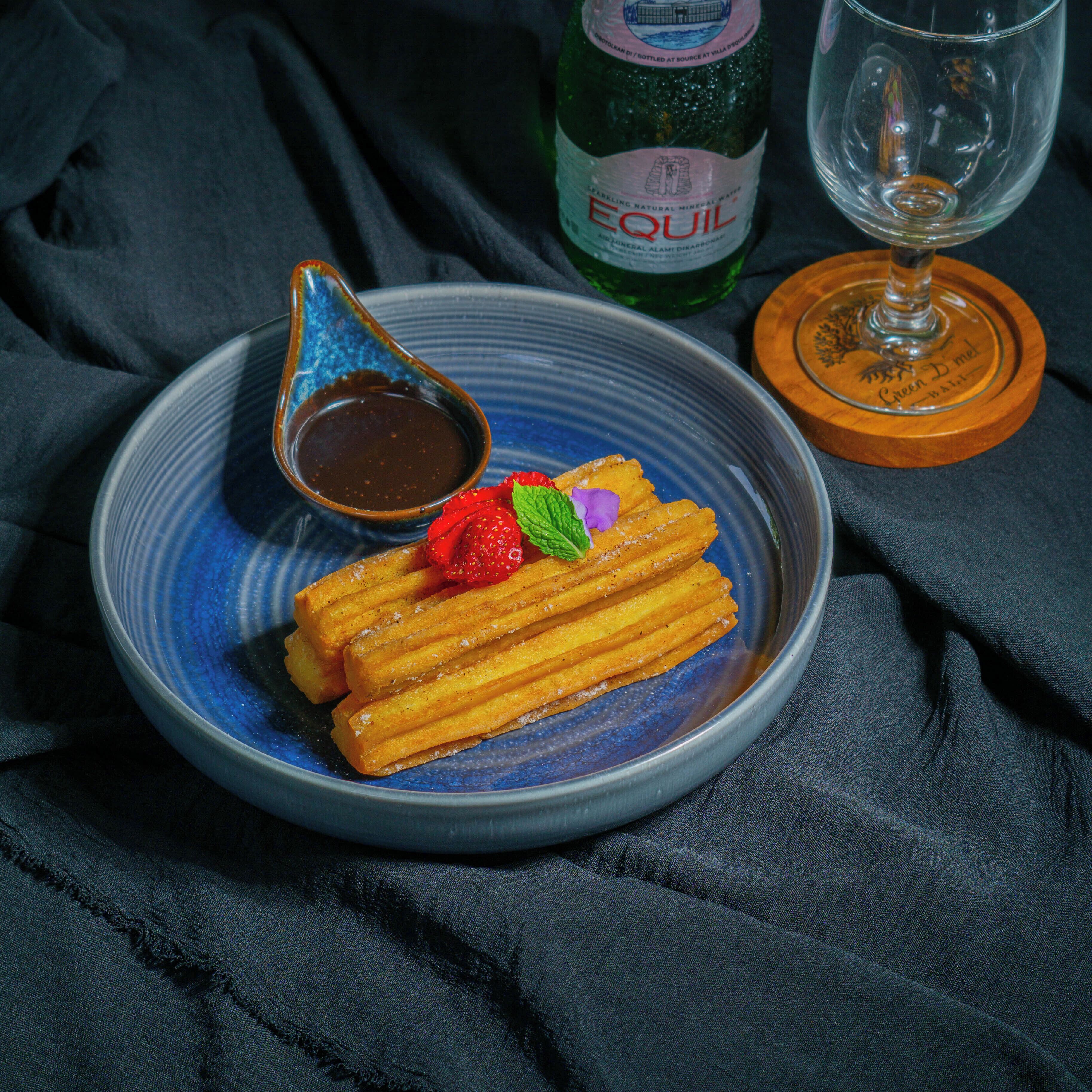 Ein Teller mit Churros, einer Tasse Schokoladensauce, einer Erdbeere und Minze auf einem blauen Teller.