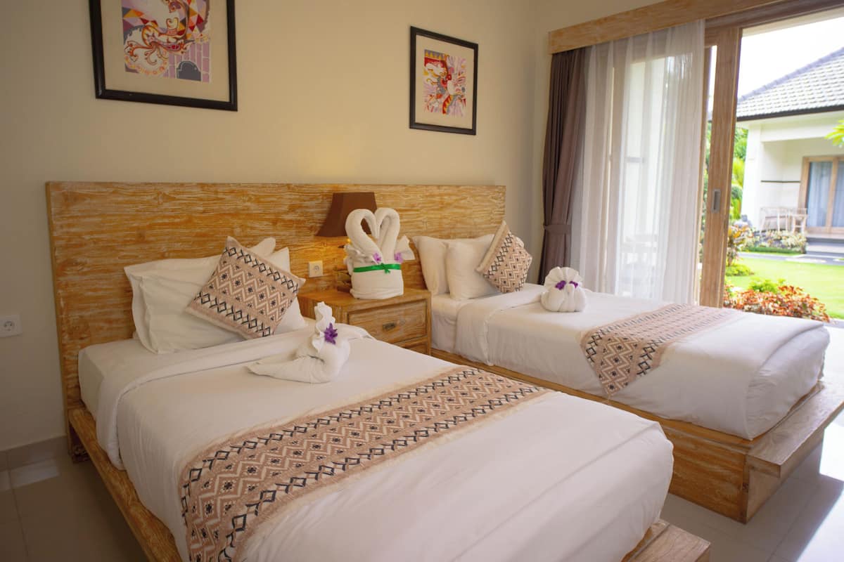 A hotel room with two beds, each with a wooden headboard, white linens, and decorative swan-shaped towels. There are framed artworks on the walls, and a view of a garden through a window.
