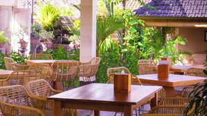 An outdoor seating area with wicker chairs and tables, surrounded by lush greenery and a tiled roof structure. - Green D'mel Bali (Nusa Dua)
