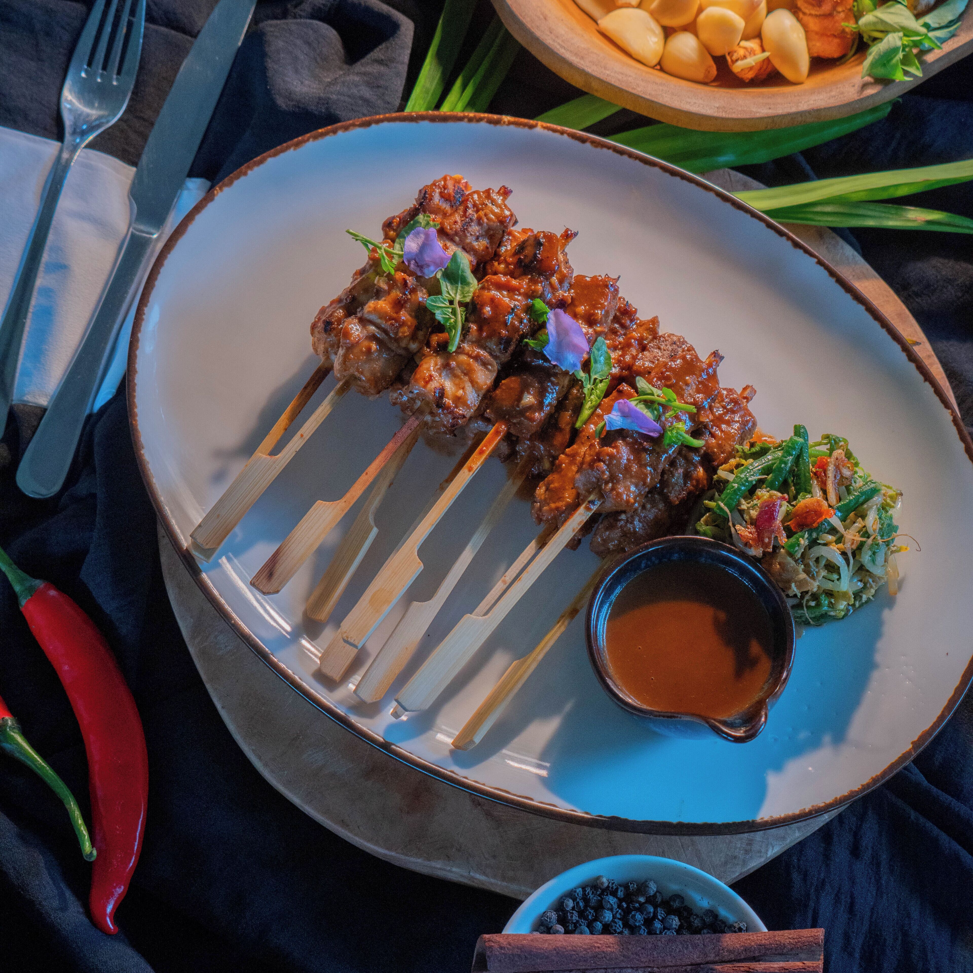 A plate of skewered meat garnished with herbs, accompanied by a side of vegetables and a small bowl of sauce.