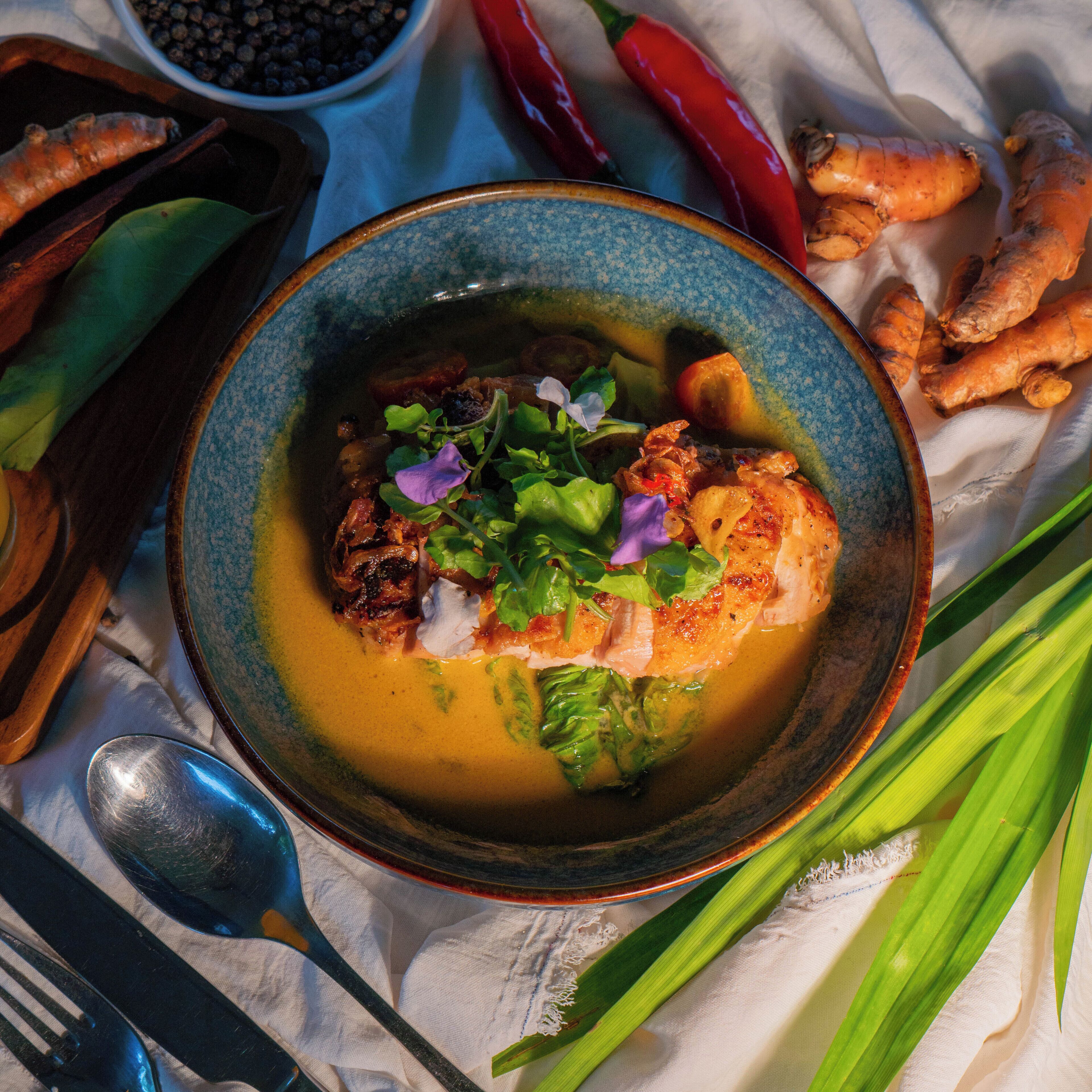 A bowl of soup with vegetables and meat, surrounded by various food items and utensils.