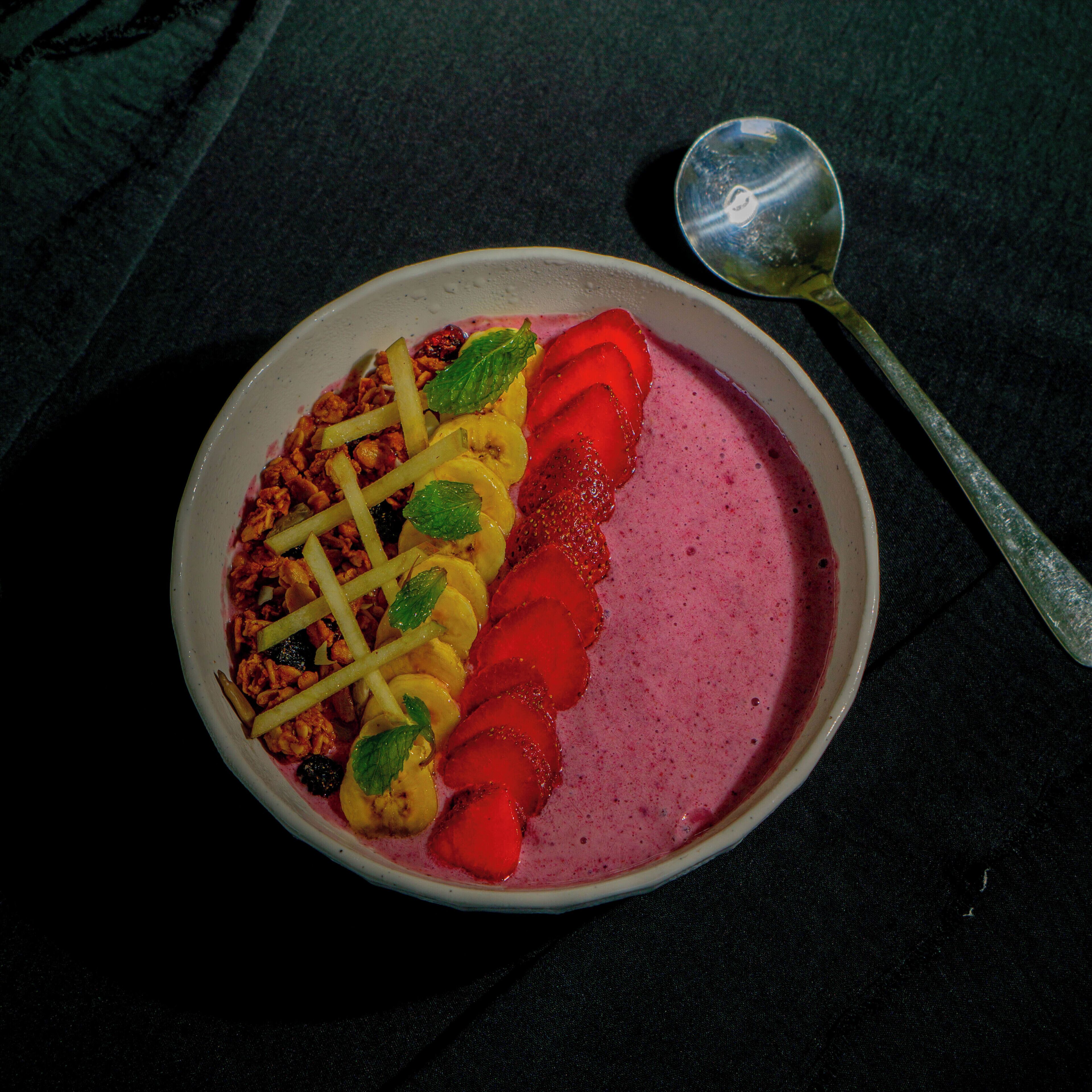 A bowl of food with sliced fruits and a spoon on a dark surface.