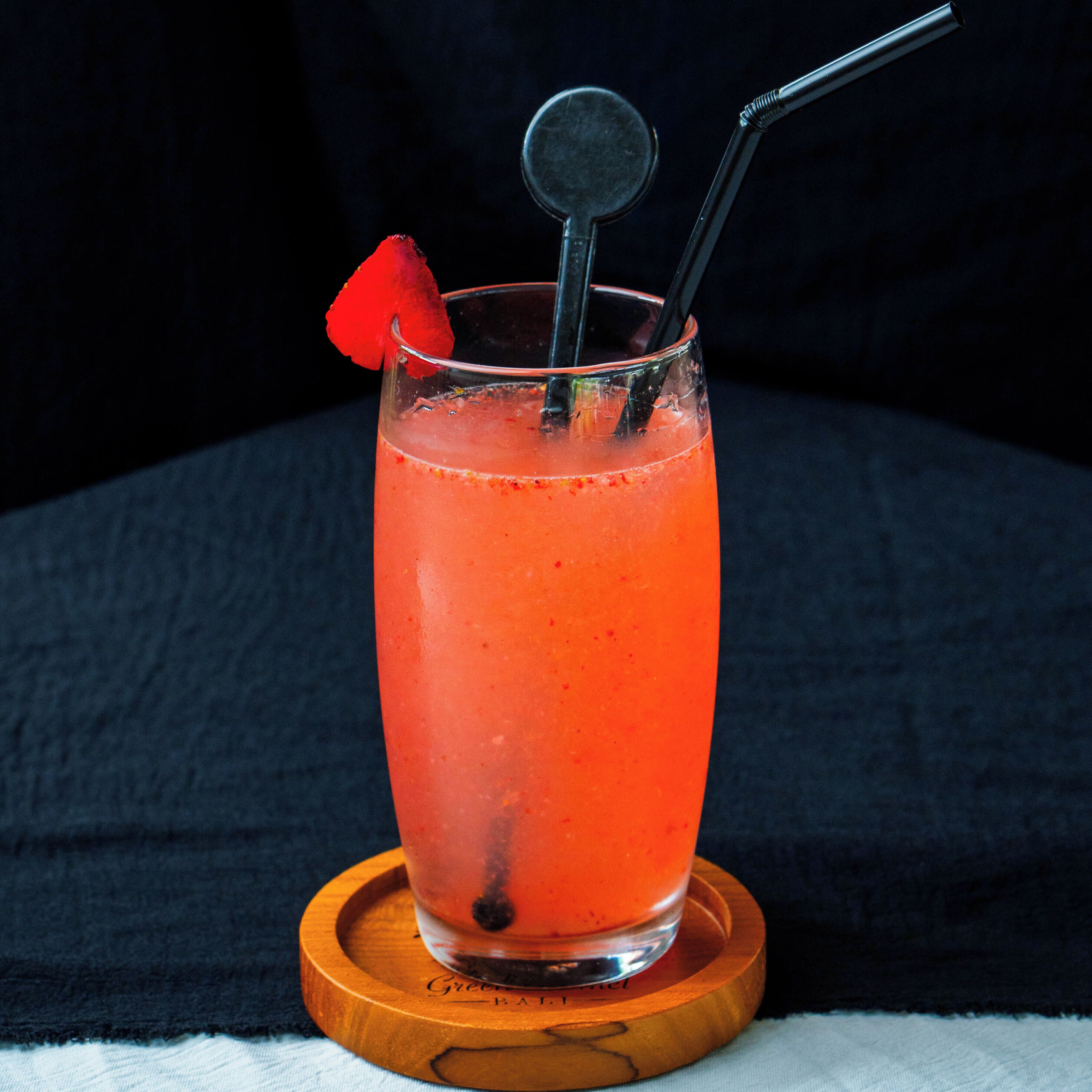 A tall glass with a red drink, a black straw, and a cocktail garnish on a wooden coaster.