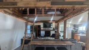 Un interior acogedor con una cama en altillo, una mesa de comedor de madera con cuatro sillas, una chimenea y una zona de cocina.