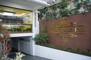 A sign for 'Heritage Inn' with a modern design, a glass enclosure with plants, and a white wall with a metal railing. - Heritage Inn (Mysore)