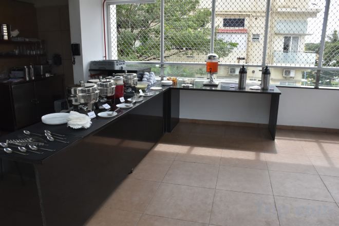 Una cocina con una encimera larga, diversos utensilios de cocina y una ventana con vistas a edificios y árboles.