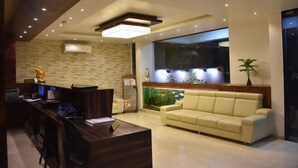 A modern reception area with a wooden reception desk, a beige sofa, and a large window overlooking a green space. - Heritage Inn (Mysore)