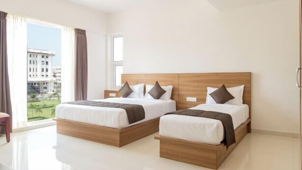 A modern hotel room with two beds, a large window with curtains, and a view of buildings outside. - Heritage Inn (Mysore)
