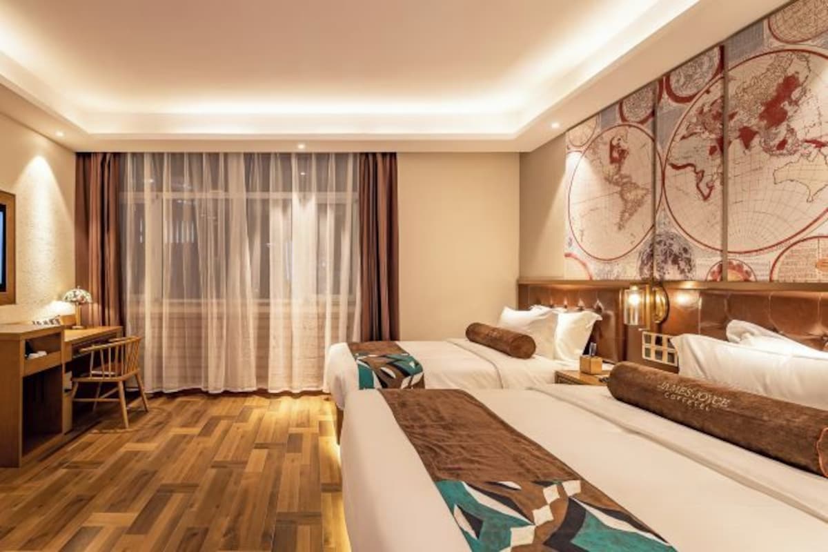 A hotel room with a wooden floor, two beds, a desk, a chair, a map on the wall, and a large window with sheer curtains.
