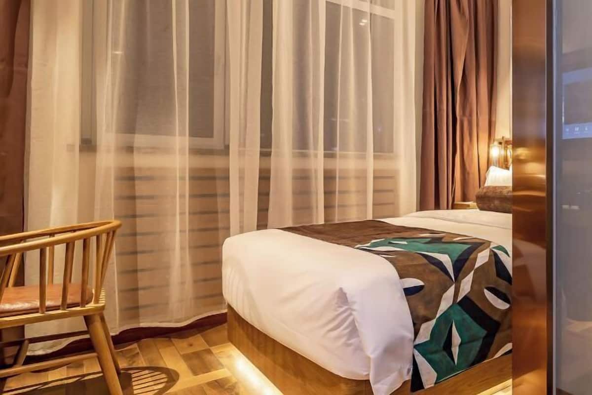 A hotel room with a bed, a wooden chair, a window with sheer curtains, and a wall-mounted lamp.