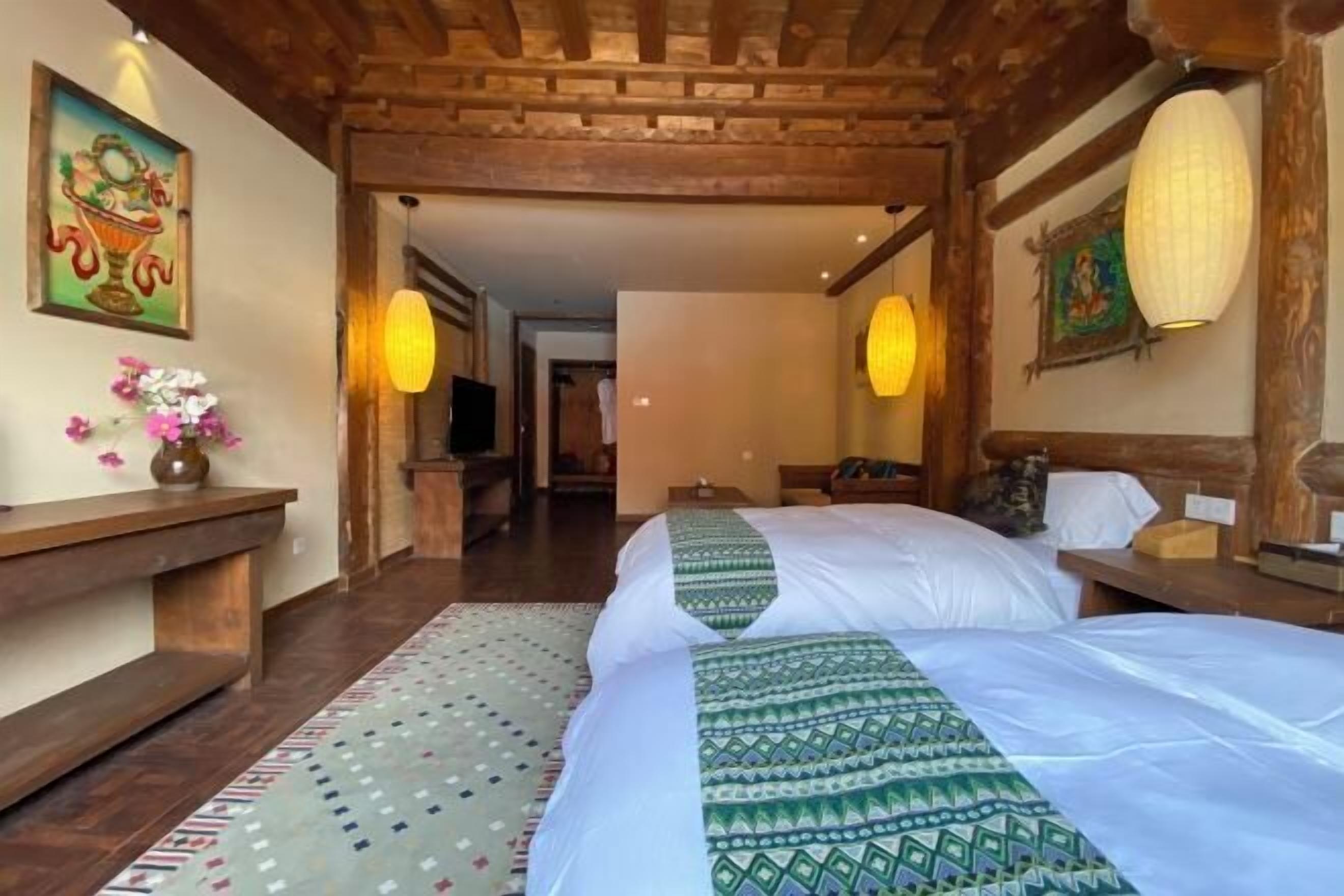 A hotel room with a wooden ceiling, two beds with patterned bedspreads, a small desk, a nightstand, a wall-mounted TV, and a decorative vase with flowers.