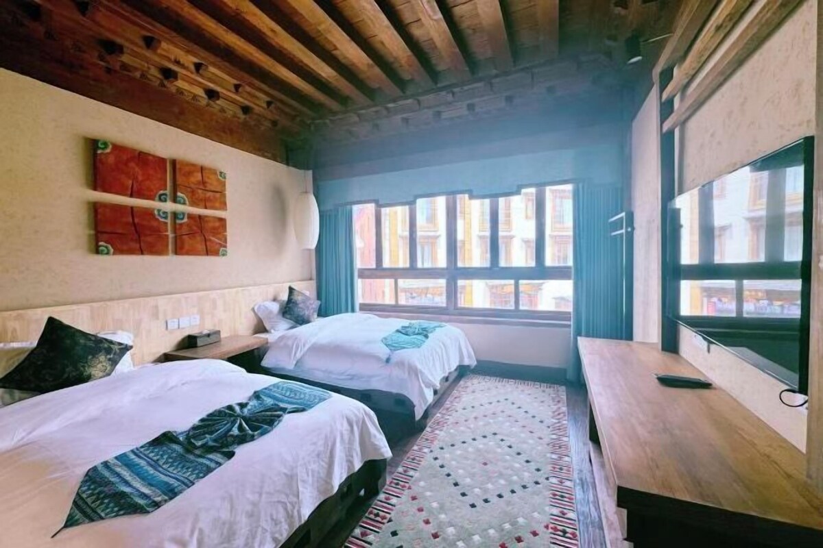 A room with two beds, a wooden ceiling, a window with curtains, and a patterned rug.