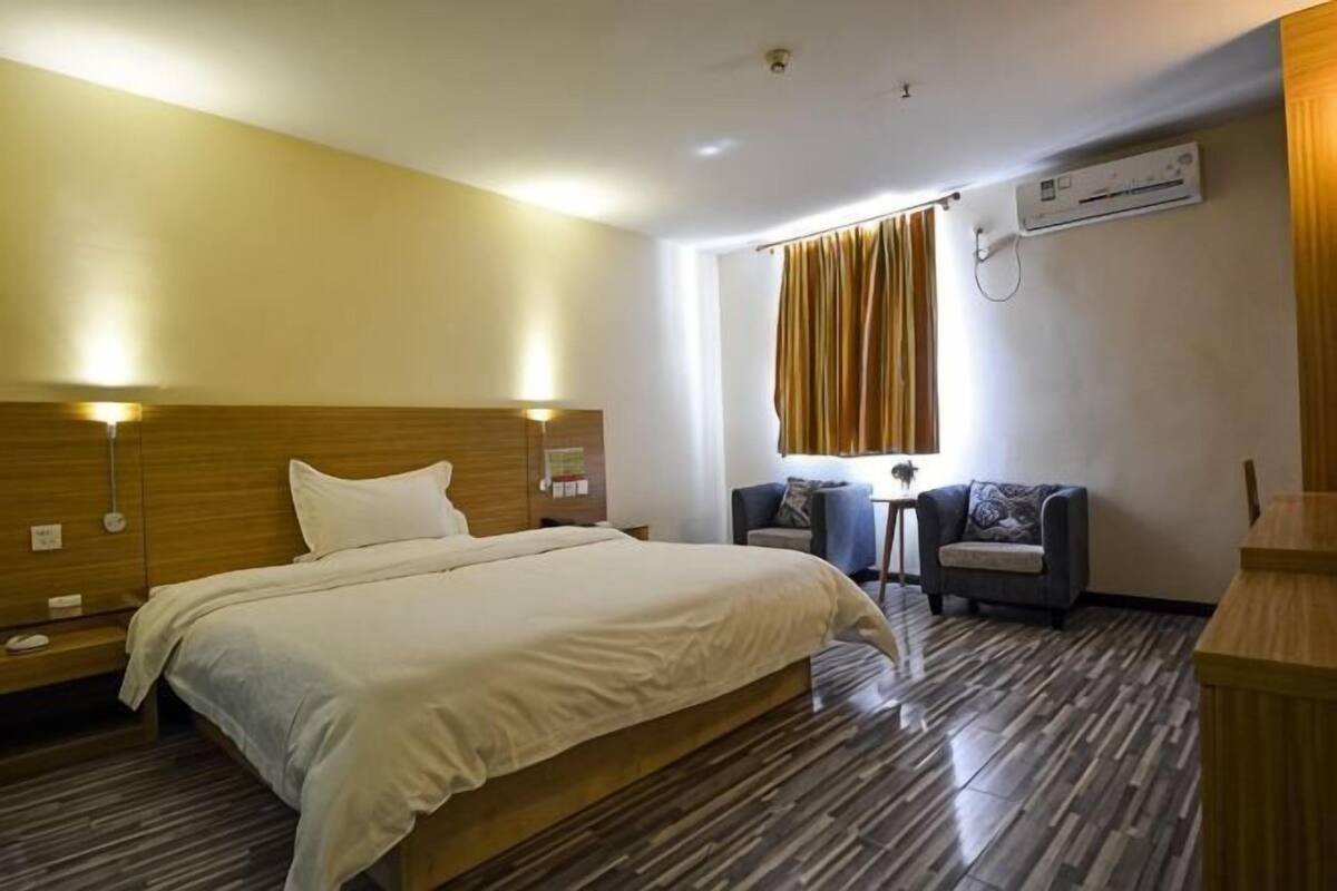 A hotel room with a large bed, a small table, a chair, a window with curtains, and an air conditioning unit.