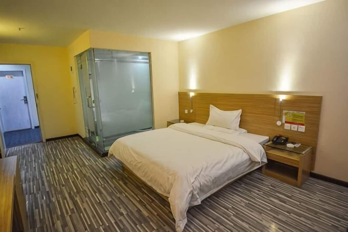 A hotel room with a bed, a nightstand, a glass shower enclosure, and a wooden headboard.
