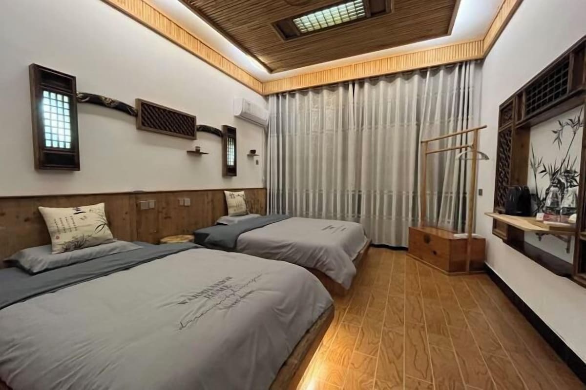 Quarto de hotel com duas camas, teto de madeira, uma janela grande com cortinas e um armário suspenso na parede.