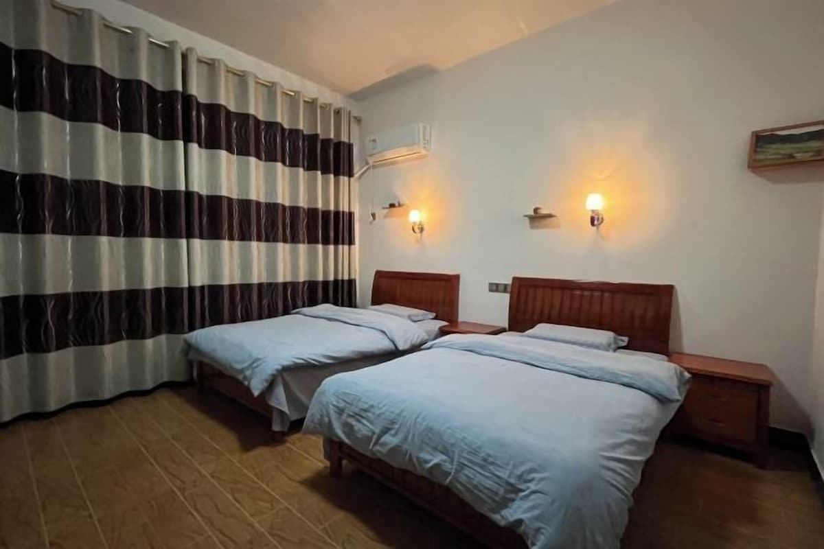 Quarto de hotel com duas camas de solteiro, cabeceiras de madeira, mesas de cabeceira, ar condicionado fixado na parede e duas luminárias de parede.
