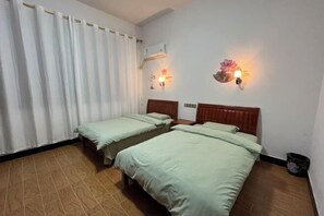 A room with two beds, a window with white curtains, an air conditioning unit, and wall-mounted light fixtures.