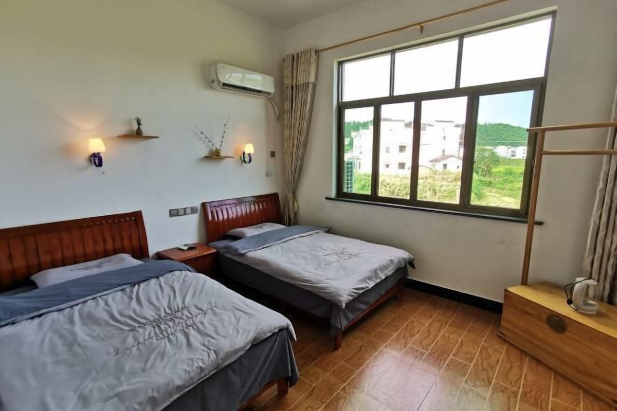 Quarto de hotel com duas camas, cabeceira de madeira, janela com vista, ar condicionado fixado na parede e mesa de cabeceira com luminária.