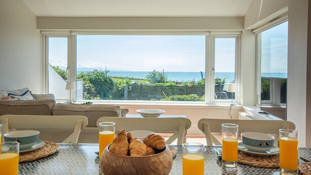 Ein Essbereich mit Meerblick, ein Tisch gedeckt mit Croissants und Orangensaft, und eine Couch im Hintergrund.