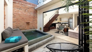 A modern living space with a brick wall, a small pool, a sofa with a blue towel, a dining area, and a kitchen.