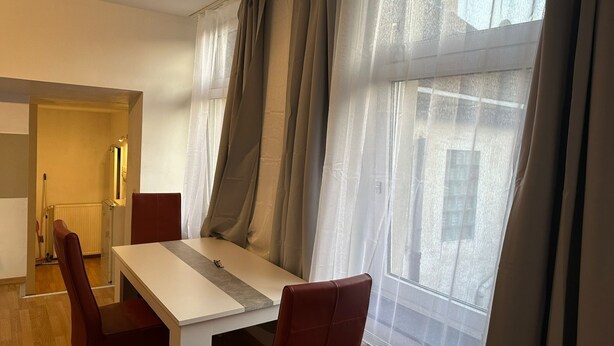 A modern dining area with a white table and red chairs, large windows with sheer curtains, and a visible radiator.
