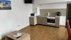 A compact kitchen with a microwave, stove, and coffee maker. A TV is mounted on the wall, and there's a painting of a beach scene.