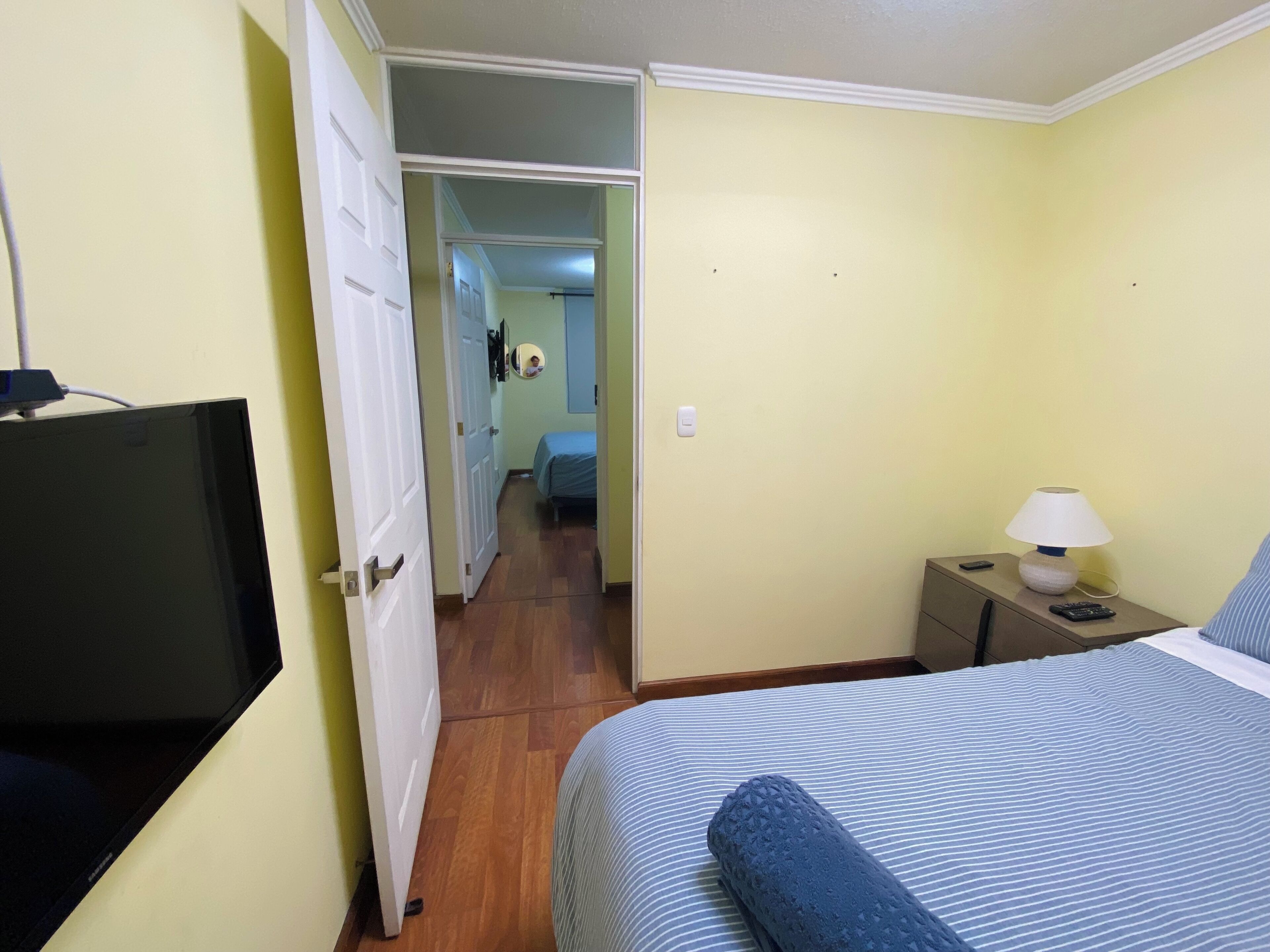 A hotel room with a bed, a TV, a bedside table with a lamp, and a door leading to another room.