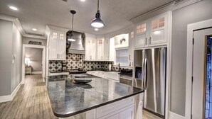 Une cuisine moderne avec des armoires blanches, un grand îlot central, des appareils en acier inoxydable et des luminaires suspendus.