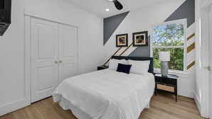 Un dormitorio con cama, mesitas de noche, ventilador de techo y ventana con vista a árboles.