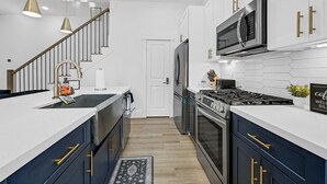 Una cocina moderna con gabinetes blancos, un refrigerador de acero inoxidable, un microondas y una estufa a gas. Hay una escalera de madera con un tapete blanco y negro que lleva a una puerta blanca.