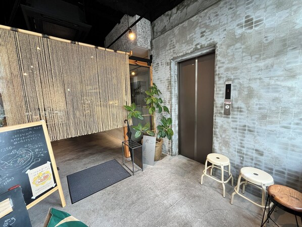 A modern interior with a textured wall, a wooden partition, a potted plant, a chalkboard menu, and a small seating area with stools. - HOTEL SEN (Matsue)