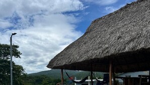 A swimming pool with a thatched roof structure, surrounded by a landscaped area with chairs and a view of green hills. - Rancho Agua Fría (Jucuarán)