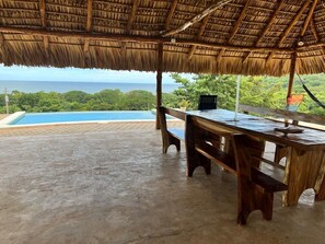 A covered outdoor area with a concrete floor, a wooden table, and benches. A pool is visible in the background. - Rancho Agua Fría (Jucuarán)