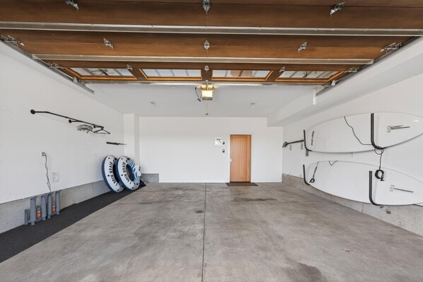 A spacious garage with concrete floors, wooden beams, and a skylight. There are two white surfboards mounted on the wall and two inflatable rings against the wall.