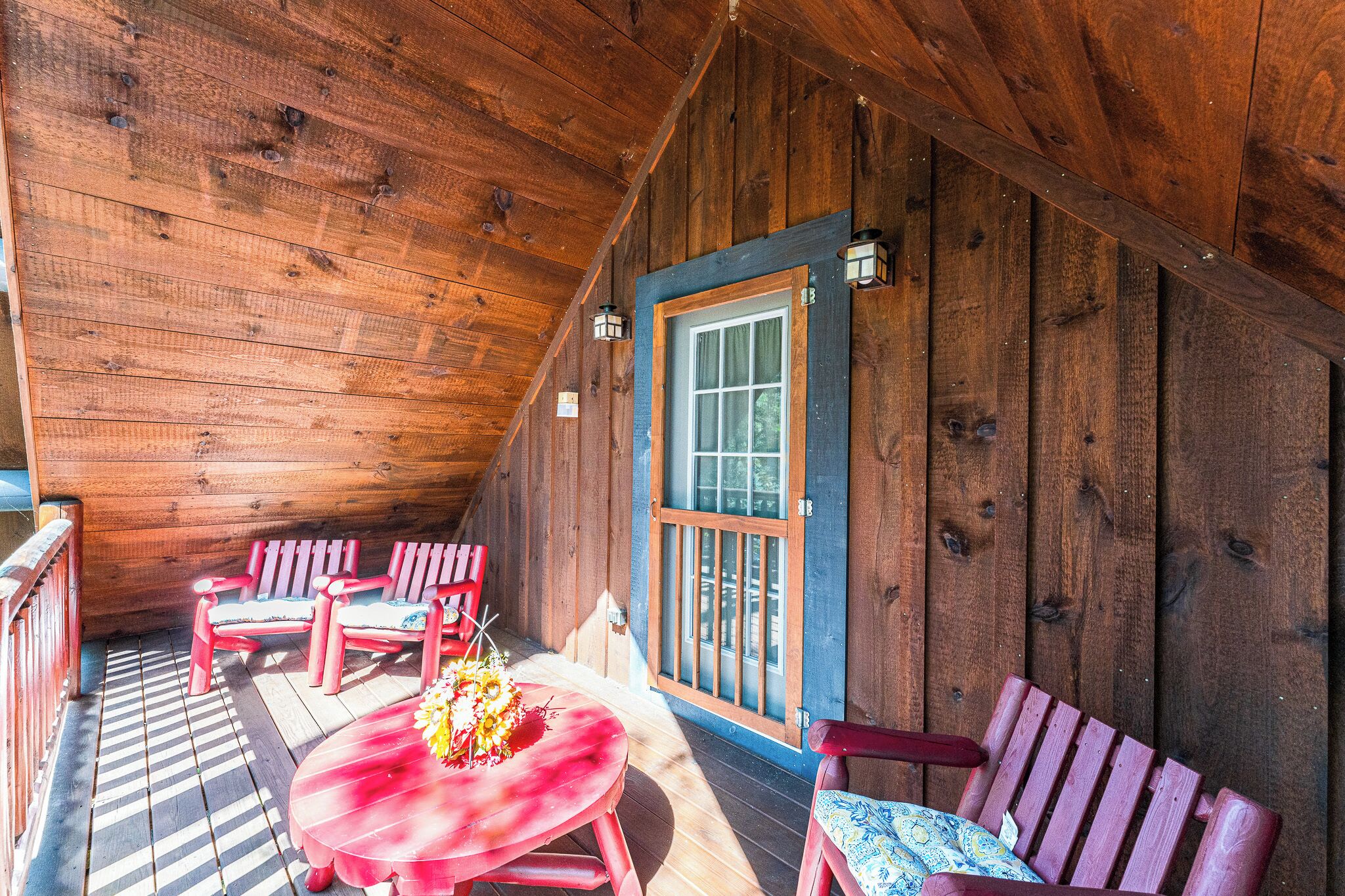 Una terraza de madera con sillas rojas y una mesa, una ventana con marco azul, y techo de madera.